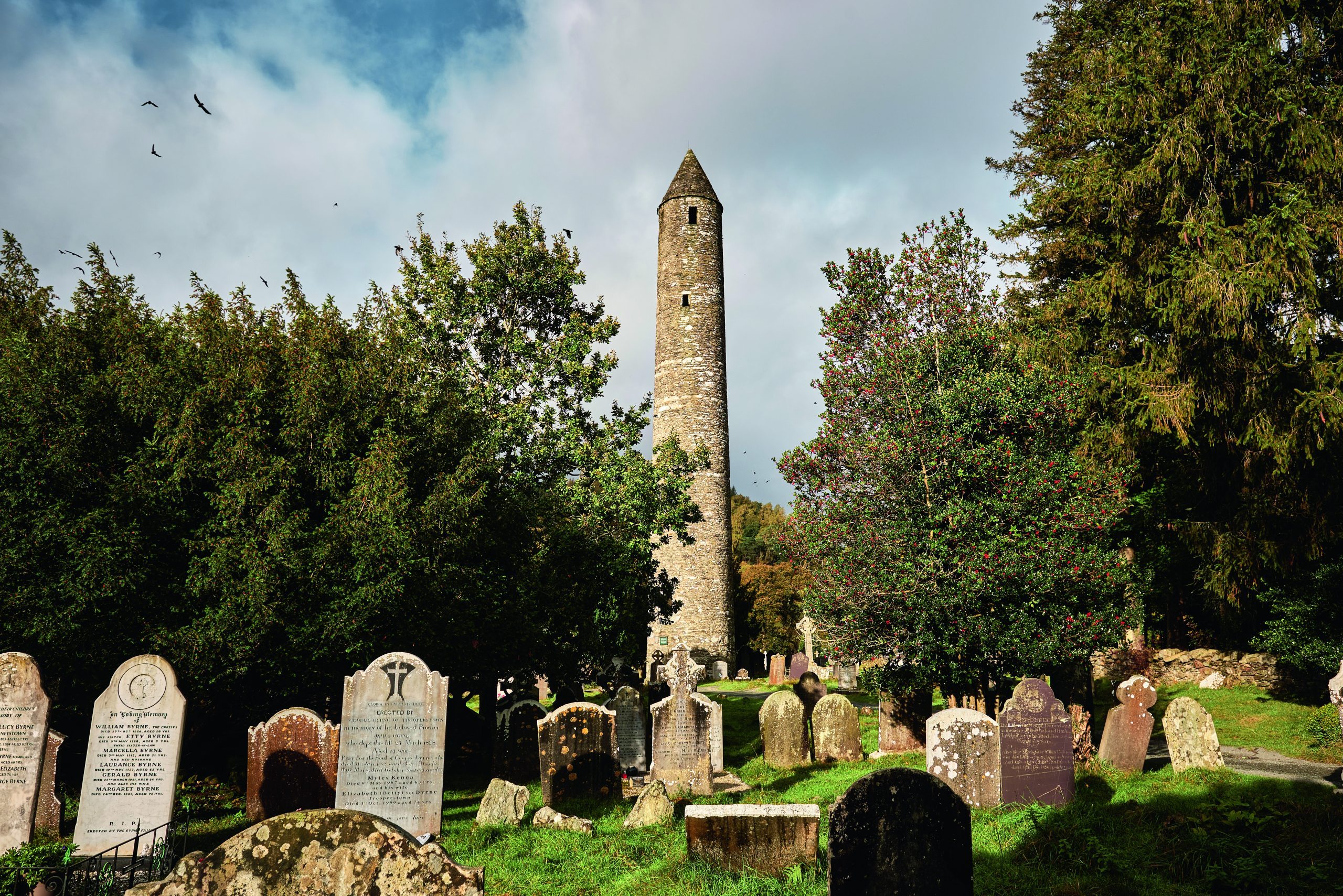 Glendalough, Ireland; 17th of October 2024. A scenic view of the iconic round tower and ancient gravestones at Glendalough, Ireland, surrounded by trees and nature, capturing Irish medieval heritage and spiritual atmosphere.