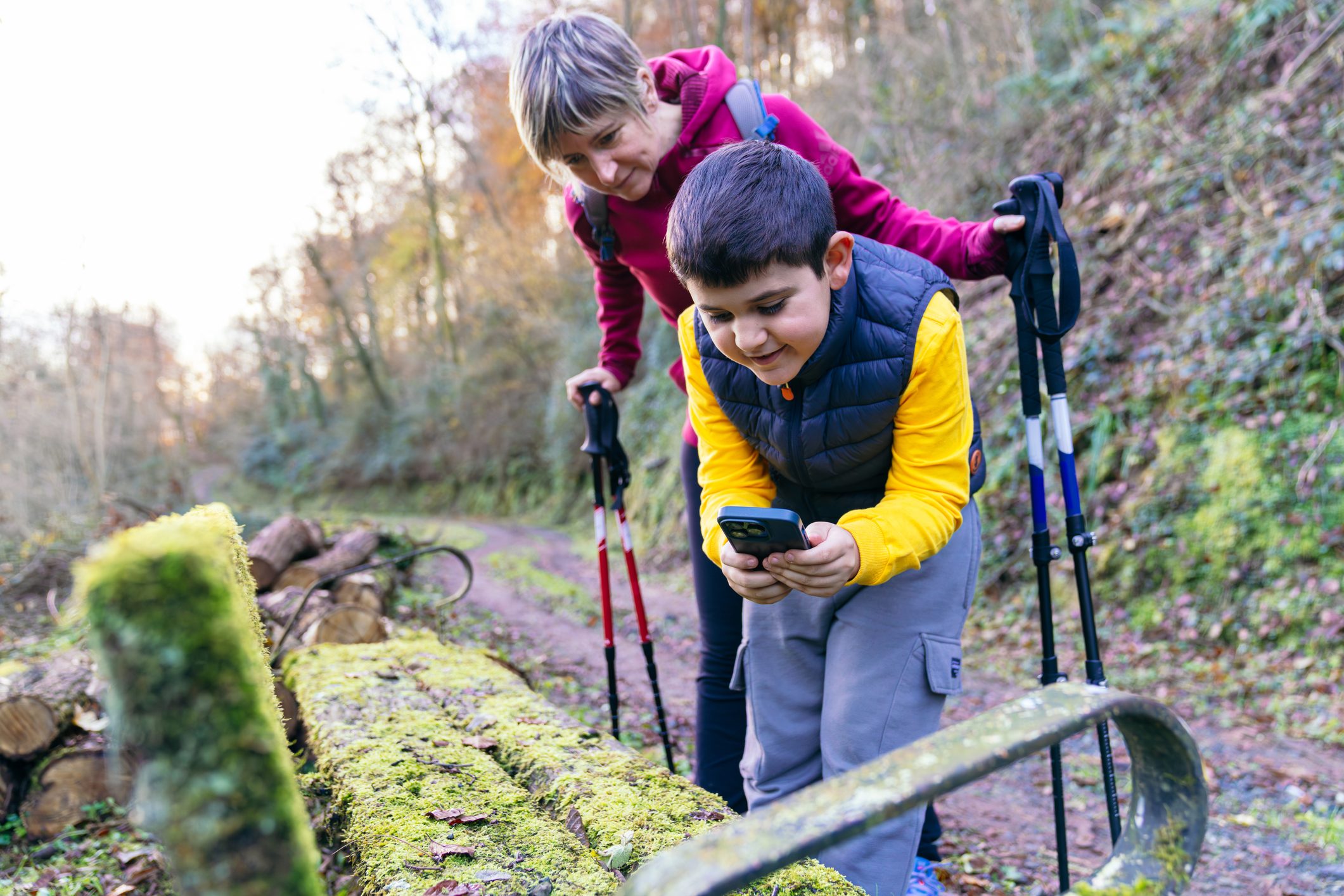 Mother and son searching for a geocache in the forest. The boy holding a smartphone and smiling, the mother leaning over him