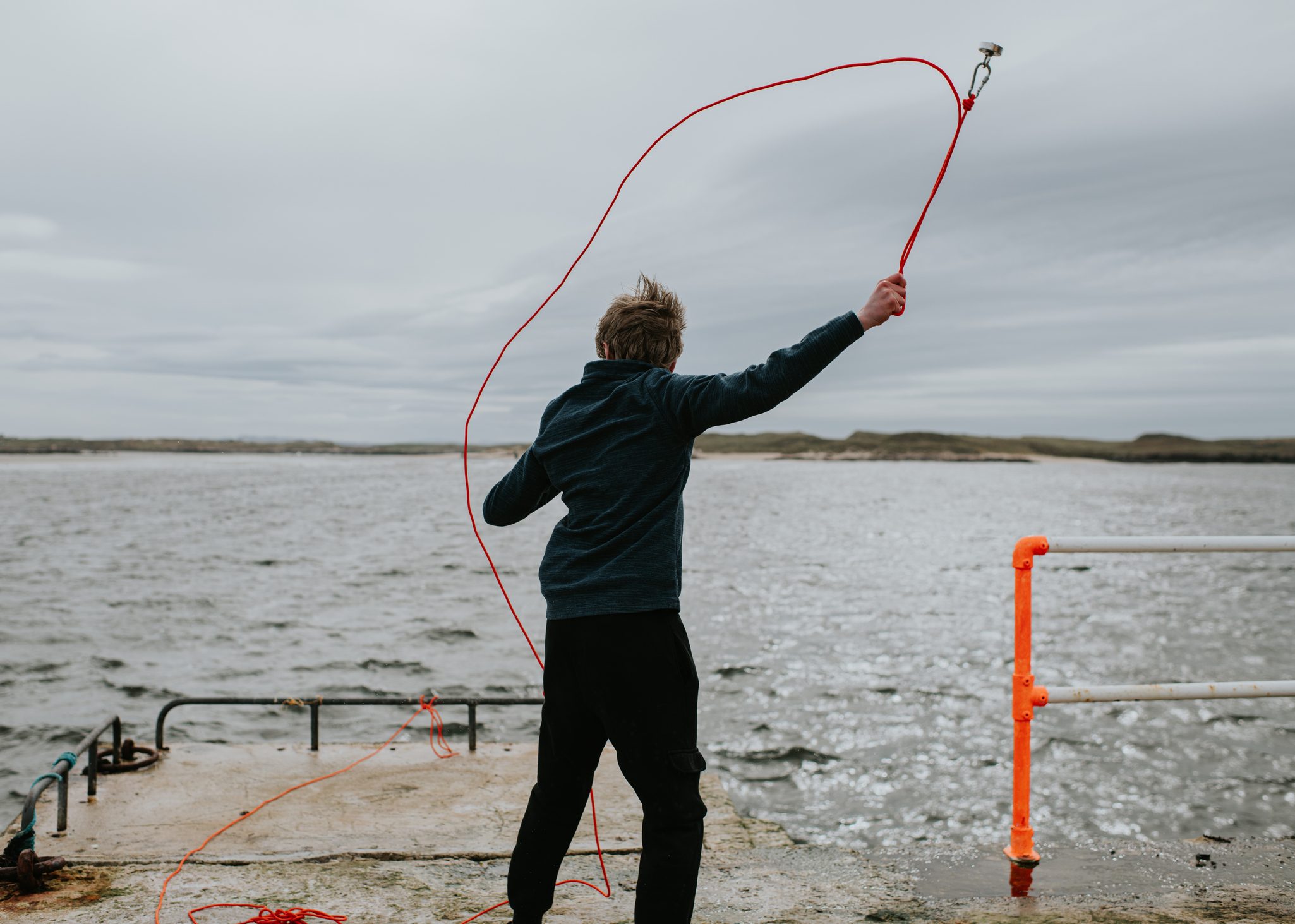 Boy standing on a concrete pier beside the sea while swinging a rope with a magnet attached for magnet fishing. The coastal landscape, cloudy sky, and open water create an adventurous outdoor scene. Suitable for concepts related to magnet fishing, outdoor recreation, coastal activities, exploration, childhood adventure, and seaside leisure.