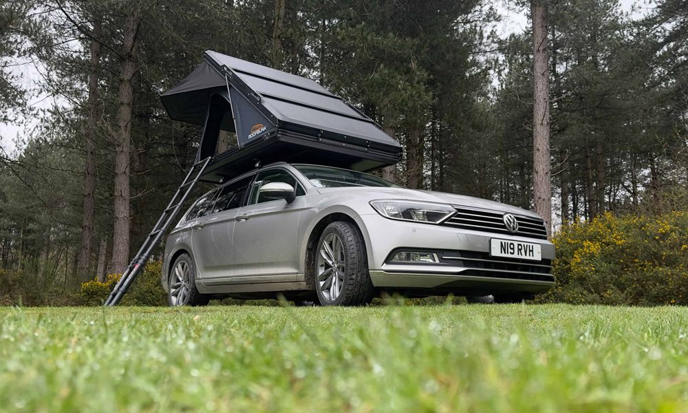 Silver car parked on grass with a black tent box expanded on the roof with a ladder coming down
