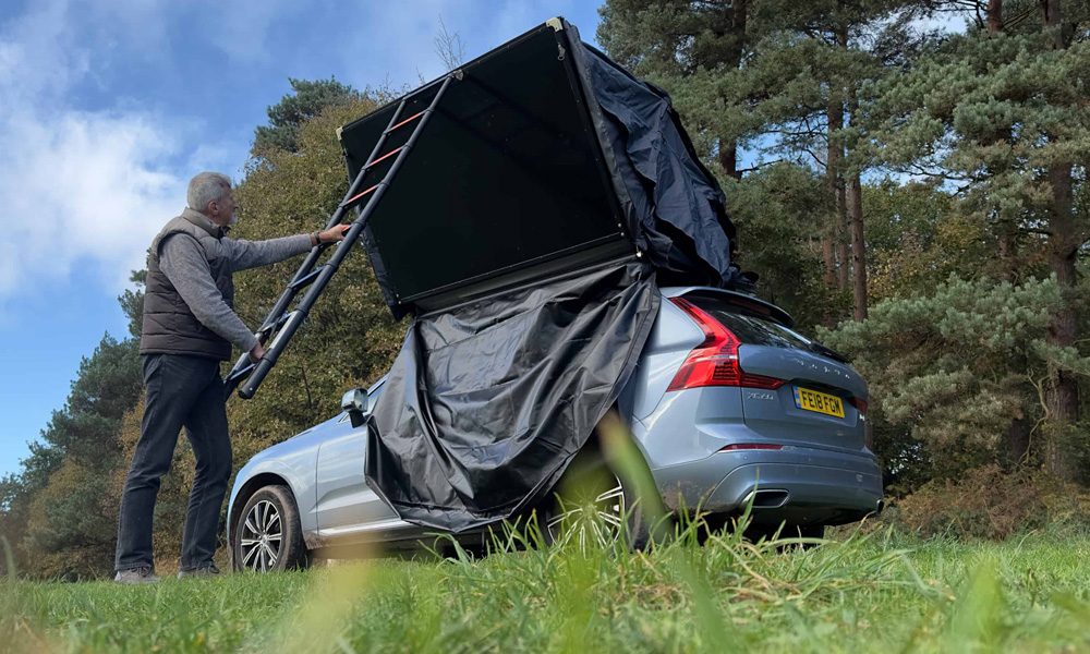 Blue car parked on grass with a grey-haired man using the ladder to expand a black tent box on the roof of the car out out