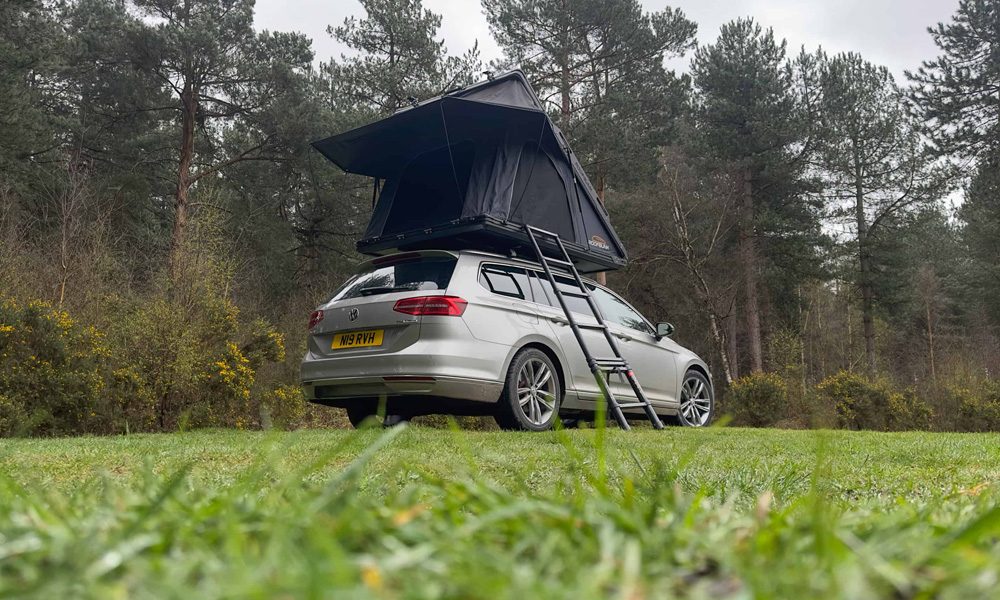 Silver car parked on grass with a black tent box on the roof and a ladder coming down to the ground