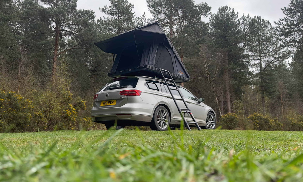 Silver car parked on grass with a black tent box on the roof and a ladder coming down to the ground