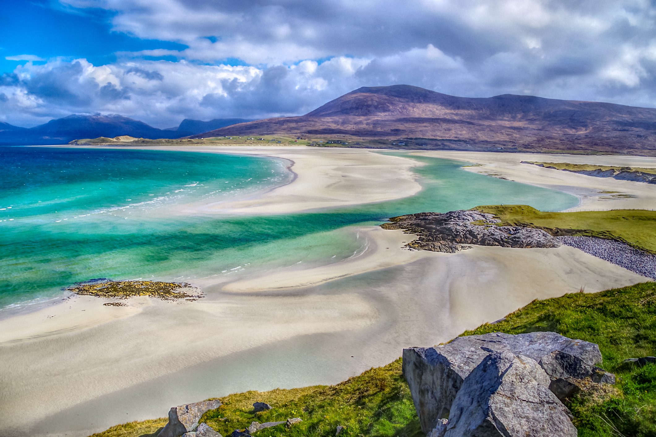 Beaches at Luskentyre on Eilean Siar in the Western Isles or Outer Hebrides (The Isle of Harris, specifically), off North West Scotland in the UK. Taken in midmorning, early May 2021 during an unusually prolonged winter. Both images show dramatic storm clouds building over the North Harris hills but these are casing shadows and unusual light where the hills meet the sea. The sand and sea at this world famous viewpoint are constantly shifting such that a new sandbank has developed and allowed the Jade Green seas to create new routes over the dazzling white sand. The two pictures are broadly similar but the second one is arguably more dramatic, given the shooting in HDR. In the first picture illustrates the deep cobalt blue sea, fading towards the hills as deep emerald but then fades further still to paler Jade in the shallowest areas. The second picture was taken in HDR and really emphasises the already vivid colours of the water, the rocks and the sand.