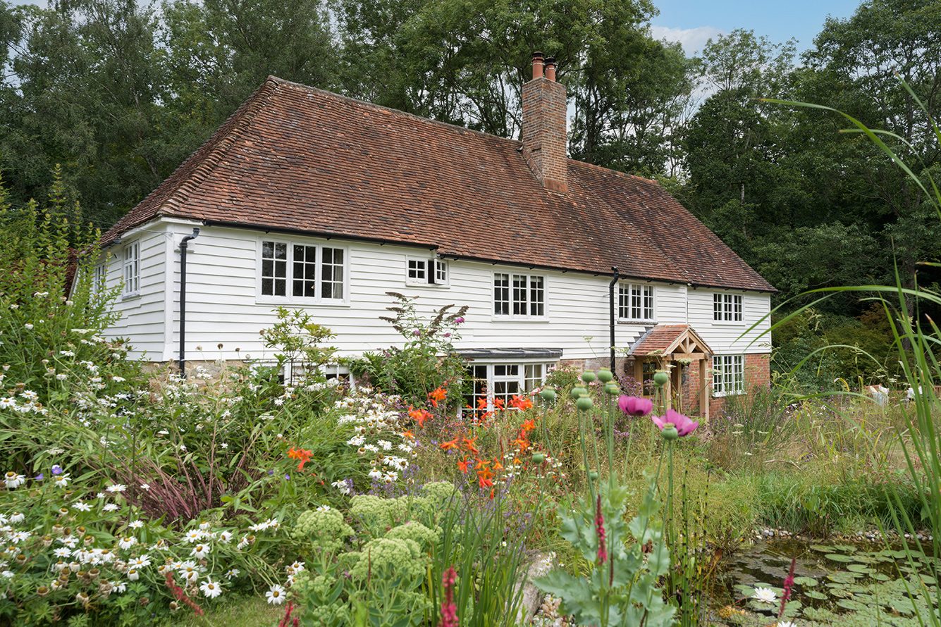 White brick cottage with terracotta roof and wooden porch surrounded by wildflowers and trees