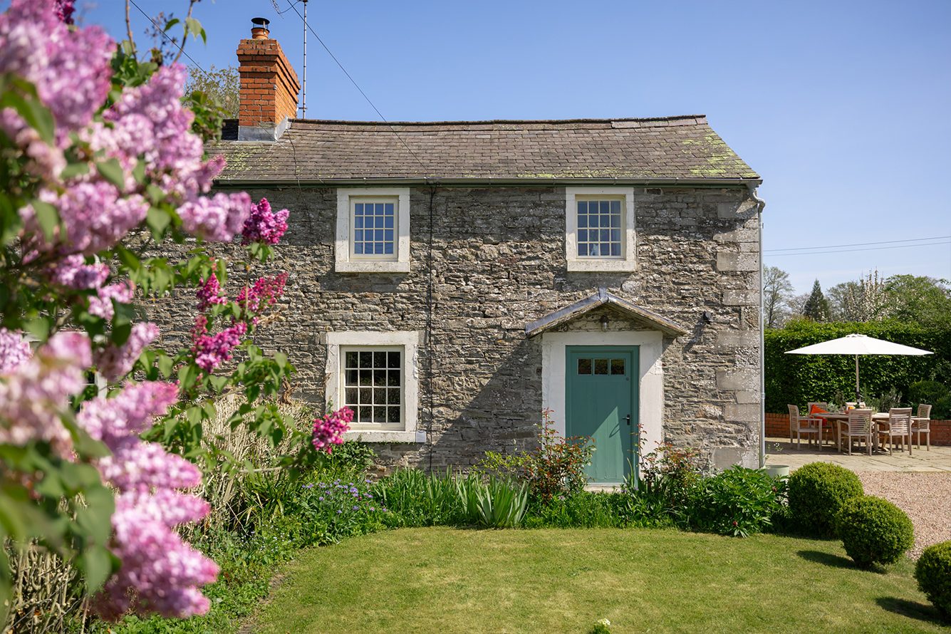 Two storey stone cottage with white window frames and green door set on a green lawn and with a pink flowering bush in the foreground