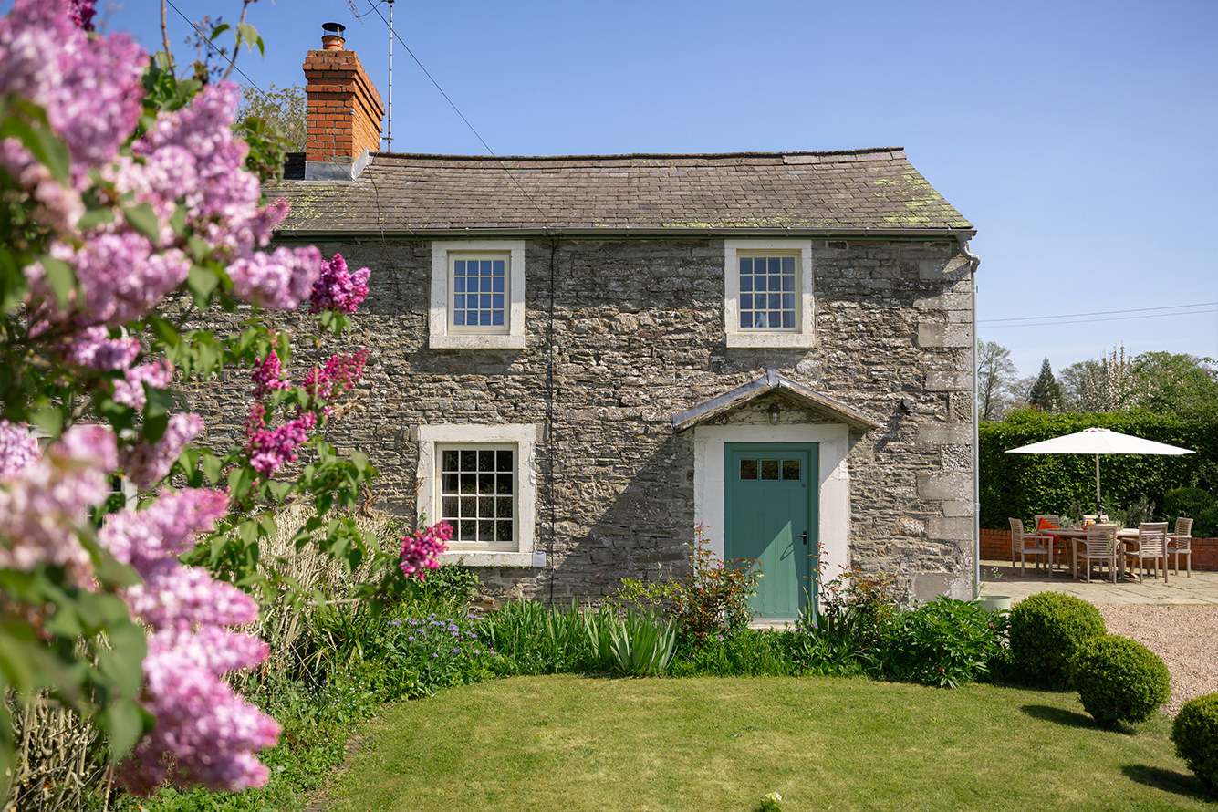 Two storey stone cottage with white window frames and green door set on a green lawn and with a pink flowering bush in the foreground