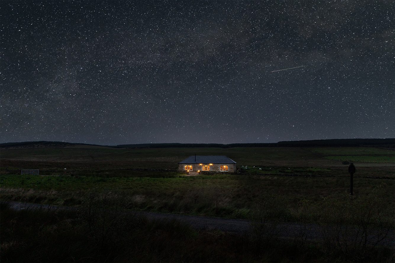 Single-storey sine cottaged with three large arched windows lit up under a star-studded night sky, set in moorland
