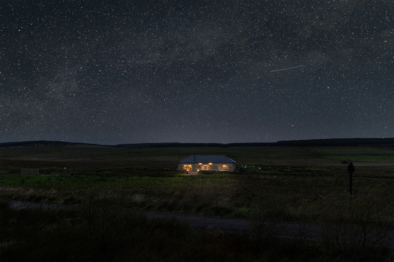 Single-storey sine cottaged with three large arched windows lit up under a star-studded night sky, set in moorland