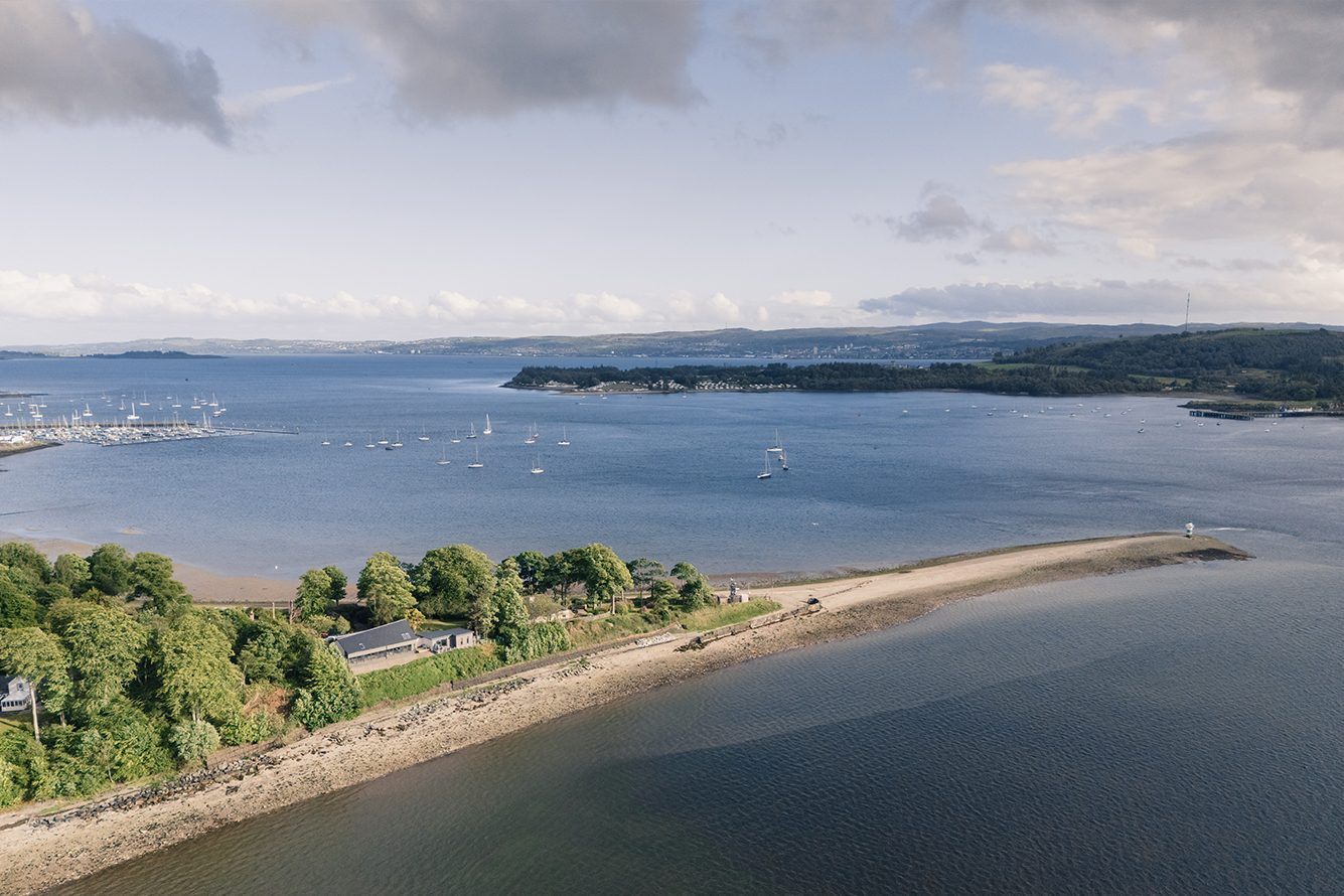 Wide angle shot of sandy spit of land stretching out into an estuary spotted with boats. There is a house visible among the trees