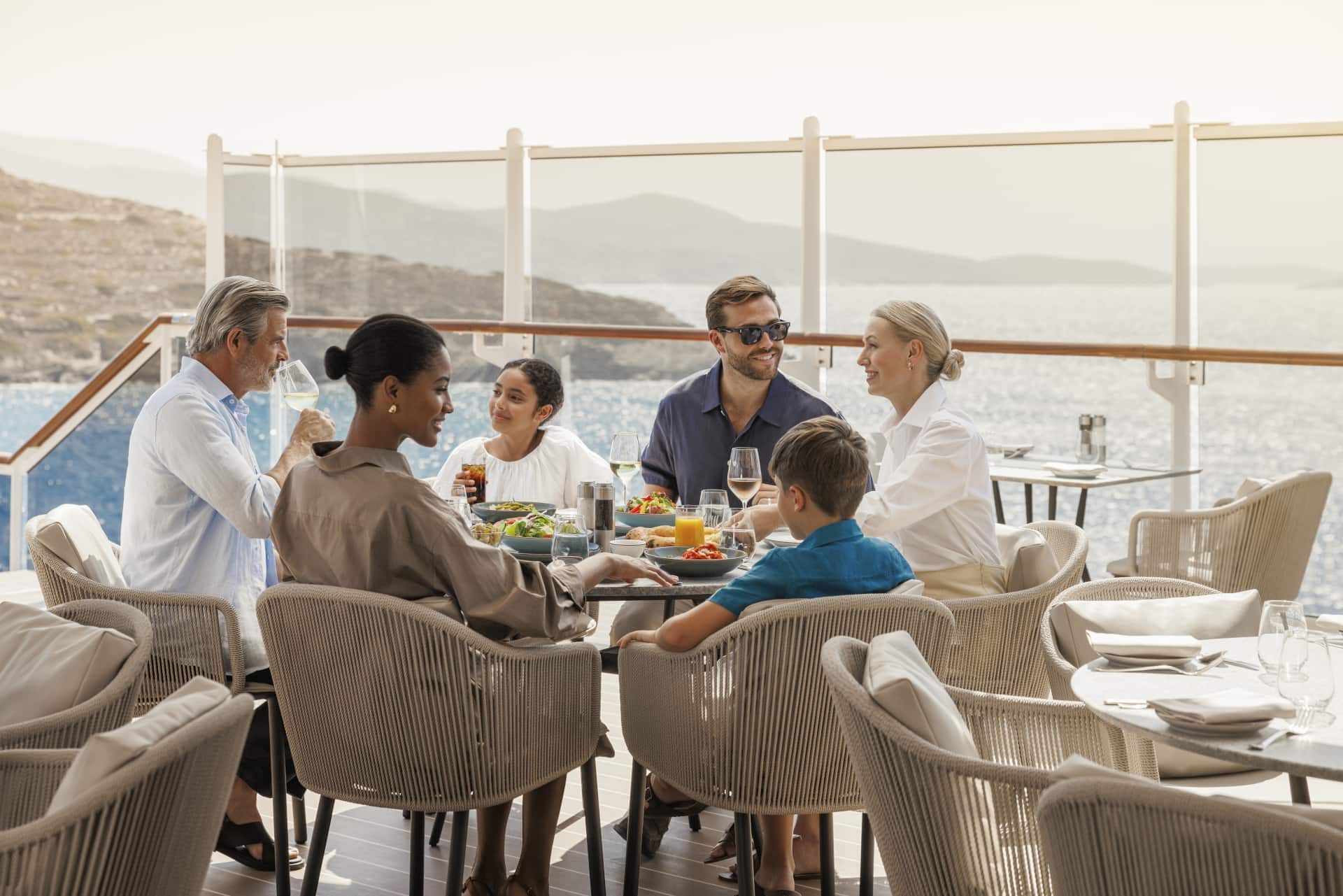 Four adults and two children sat on a sunny cruise ship deck with food and drink in front of them on the tables