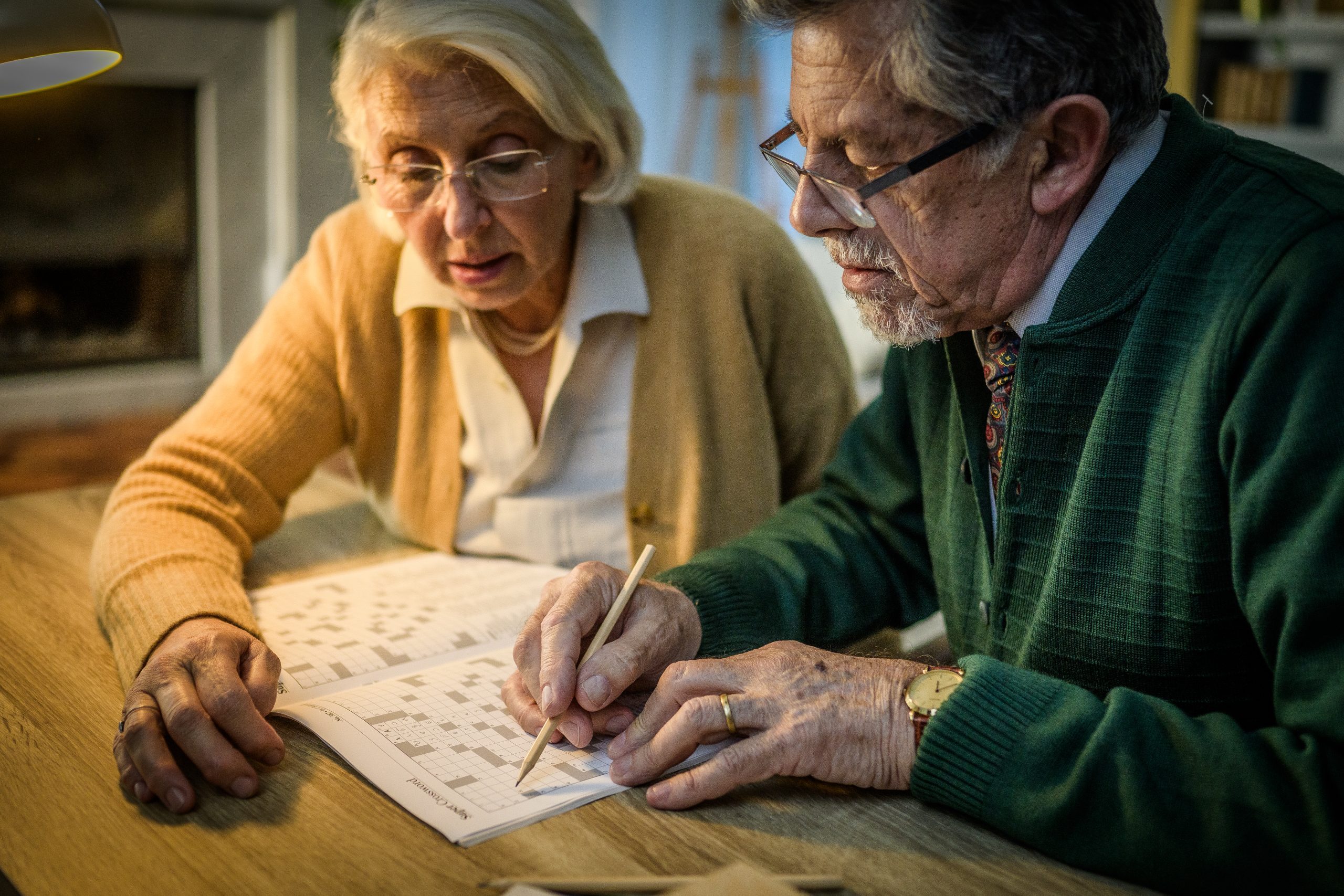 An elderly couple, a man and woman, solve a crossword puzzle together at home. They are seated at a wooden table, creating a warm and cozy atmosphere.